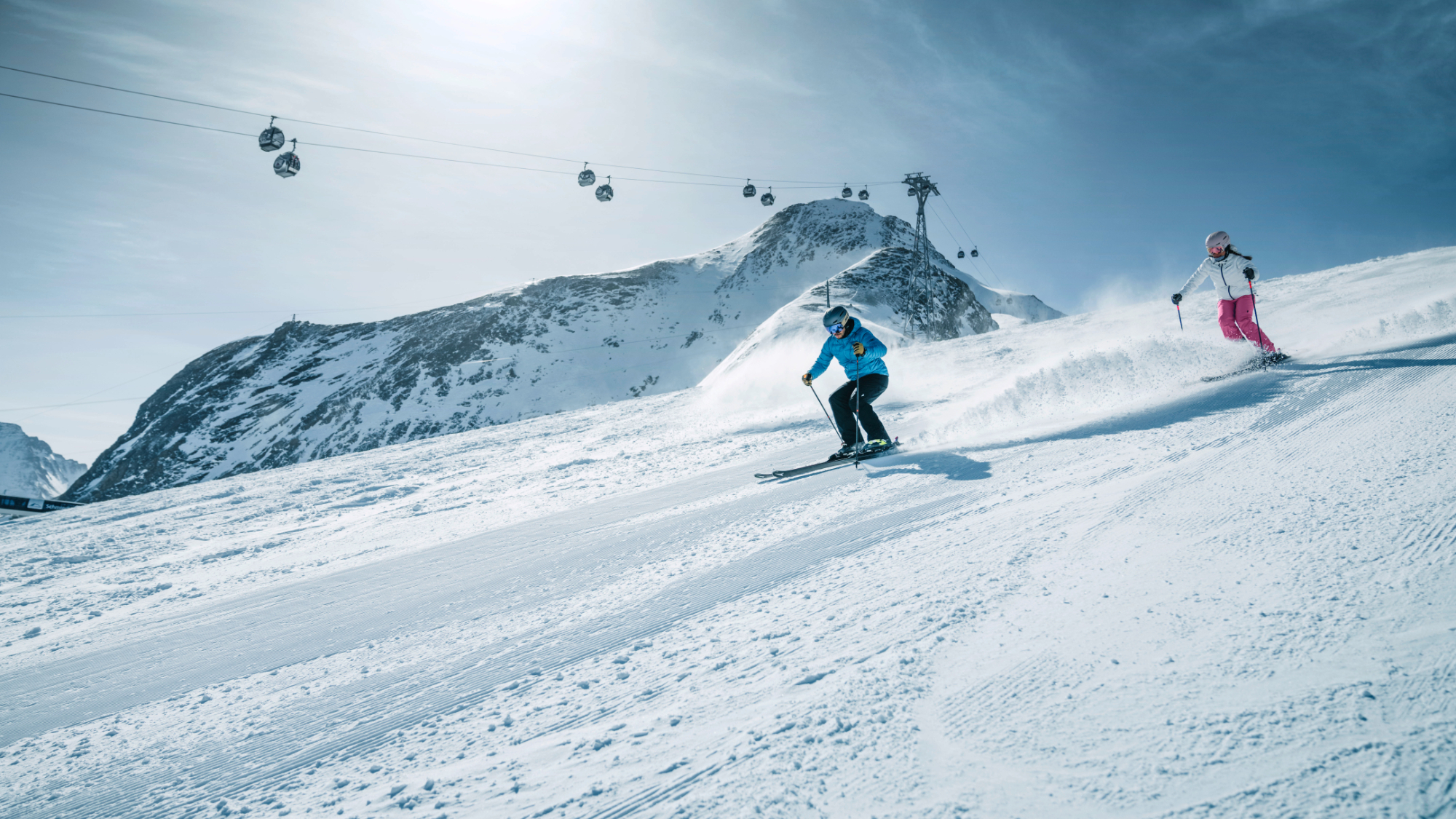 Zwei Skifahrer auf einer Piste am Kitzsteinhorn, im Hintergrund ist eine Gondelbahn und ein Gipfel sichtbar